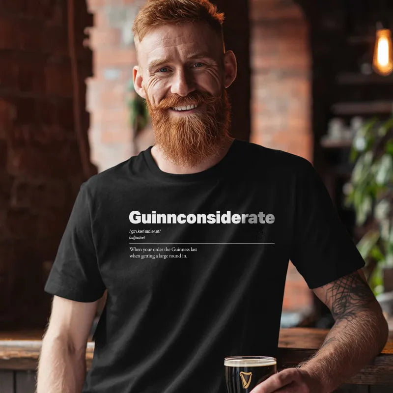 Man wearing a black t-shirt with text, saying Guinnconsiderate holding a glass of beer indoors.