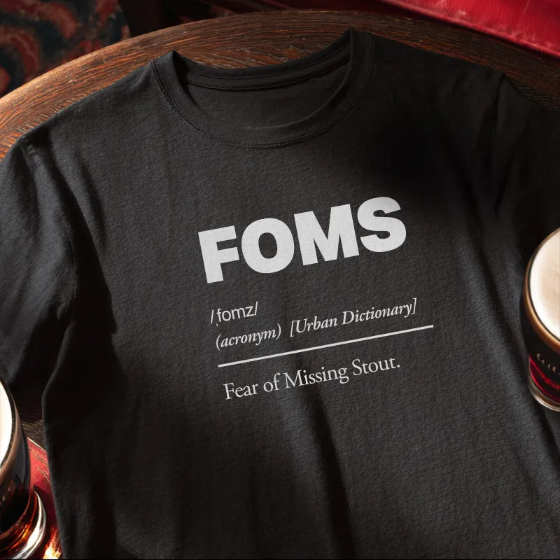 Black t-shirt with 'FOMS Fear of Missing Stout definition  placed on a wooden bar table with a glass of beer.