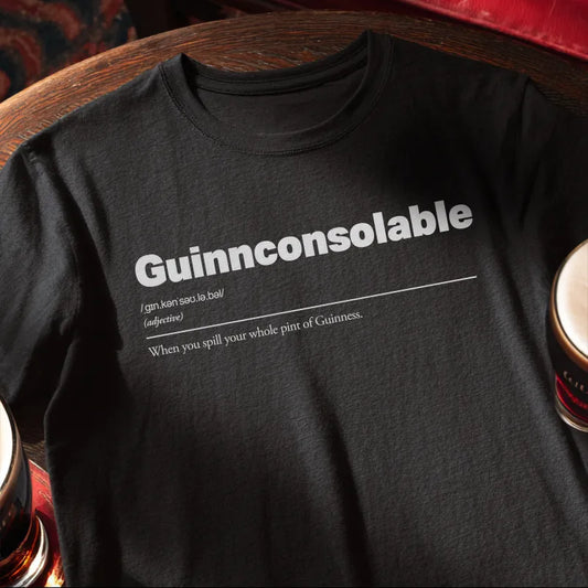 Black t-shirt with 'Guinnconsolable' text on a wooden bar table with a glass of Guinness.
