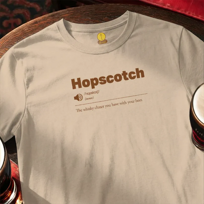 Beige t-shirt with 'Hopscotch' design on a wooden bar surface with beer glasses.