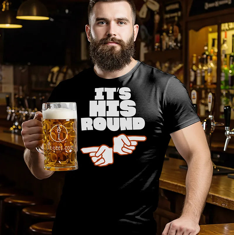 Man in a bar wearing a black beer themed  t-shirt with 'It's His Round' text, holding a beer.