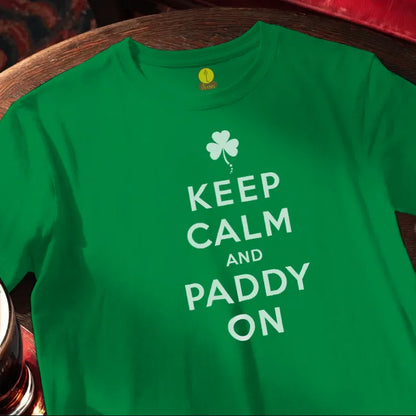 Green t-shirt with 'Keep Calm and Paddy On' text and shamrock design on a wooden bar surface.