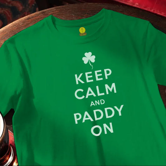 Green t-shirt with 'Keep Calm and Paddy On' text and shamrock design on a wooden bar surface.