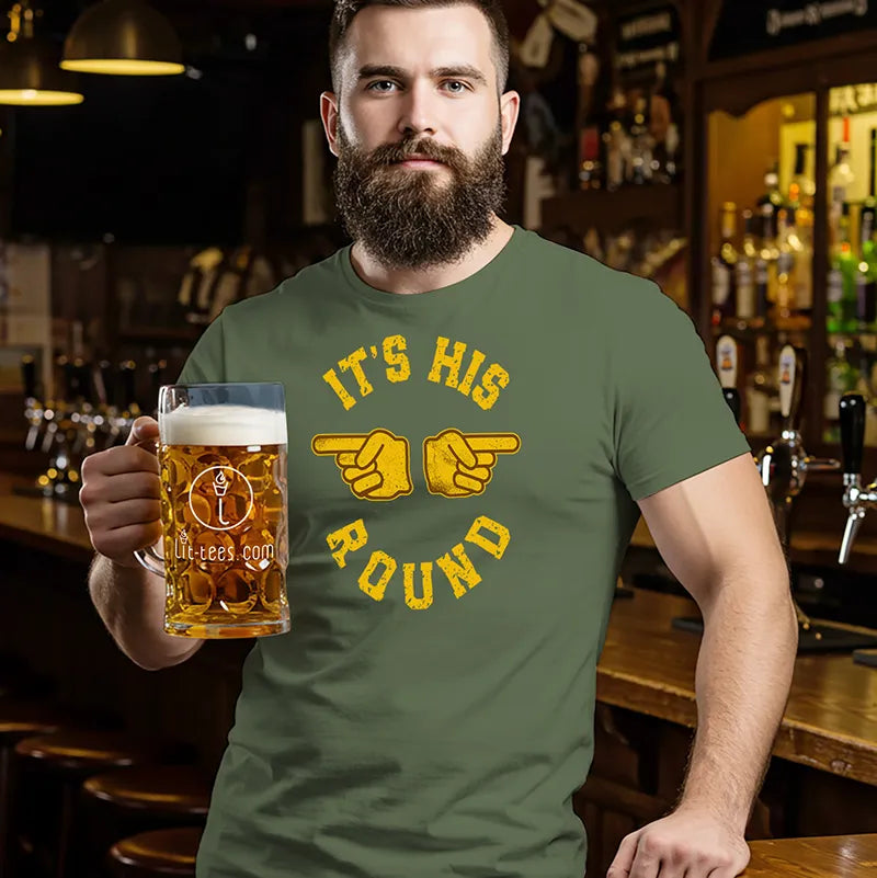 Man in a bar holding a beer wearing a military green beer themed t-shirt with yellow text saying "It's His Round'
